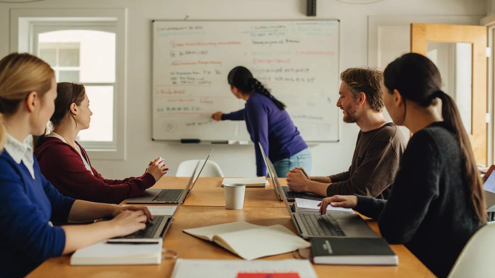 A small team in an instructor-led training session around a wooden table, one person at a whiteboard